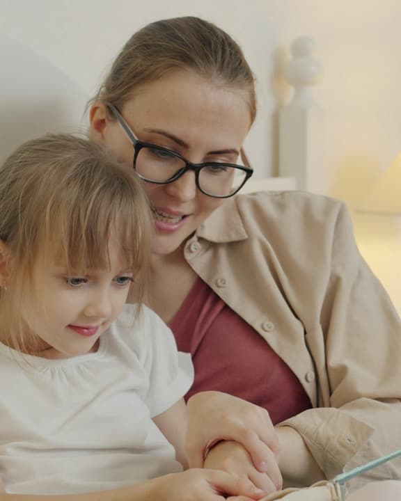 A mother and her young daughter sit close together at home, sharing a quiet moment in soft lamp light.